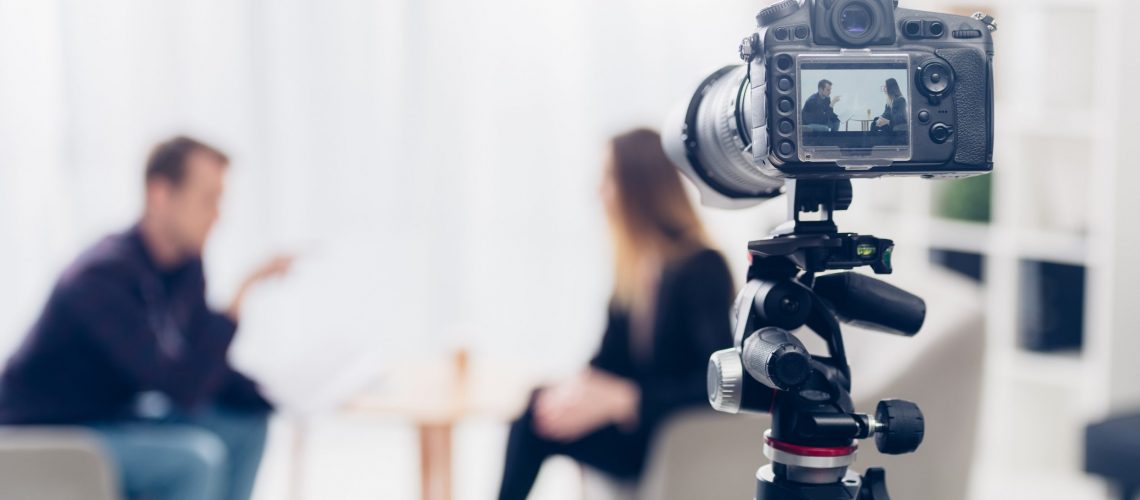 businesswoman in suit giving interview to journalist in office, camera on tripod on foreground