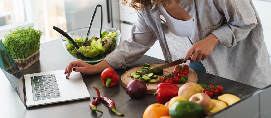 Happy young woman making a salad at the kitchen, chopping vegetables, looking at laptop computer