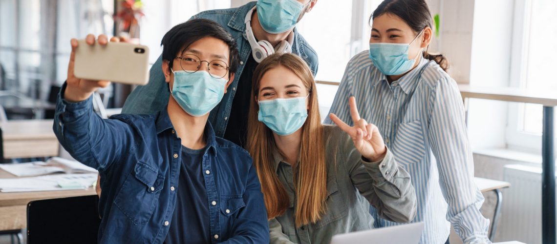 Photo of multinational cheerful students in medical masks taking selfie on cellphone at classroom