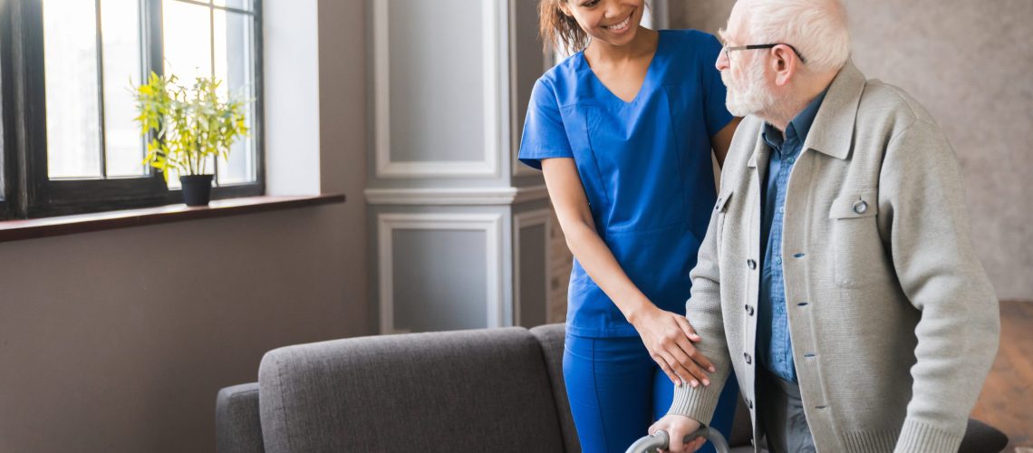 Portrait of an african young nurse helping old elderly disable man grandfather to walk using walker equipment in the bedroom. Senior patient of nursing home moving with walking frame and nurse support