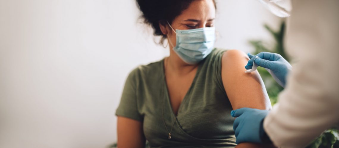 Woman wearing protective face mask getting vaccine shot at home. Female receiving covid vaccination by a healthcare worker at home.